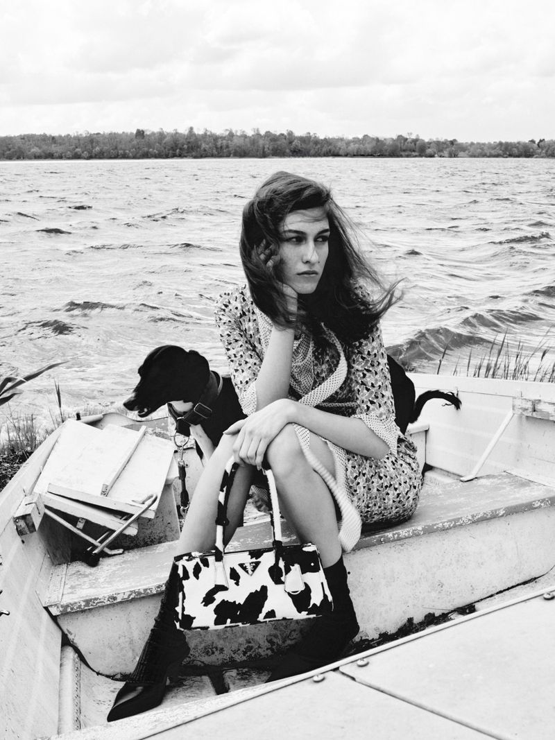 Black and white outdoor portrait of a female adult sitting in a boat with a dog, wearing a dress and boots, holding a purse.