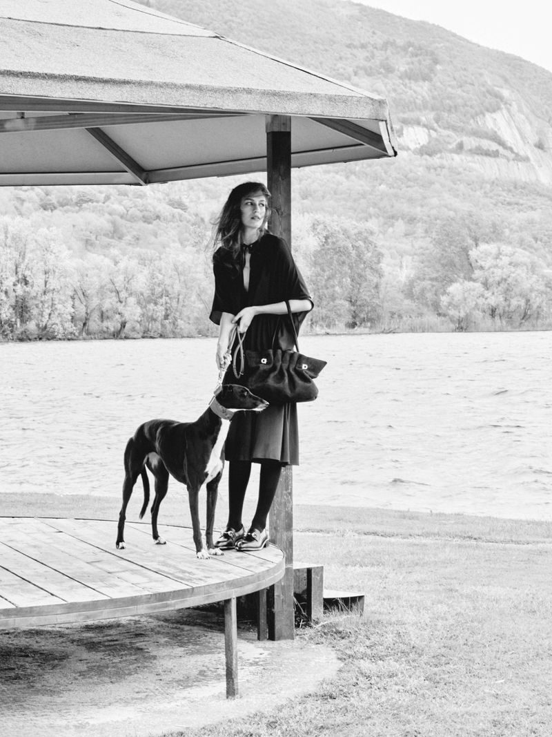Black and white outdoor shot of a woman in a dress, standing with a greyhound dog next to a lake and mountain landscape.