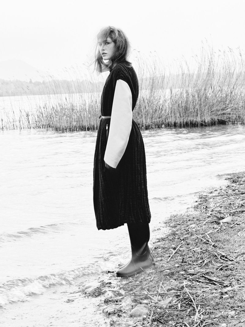 A black and white shot captures a woman in a dark dress and boots standing near water and tall grasses at the edge of a lake.