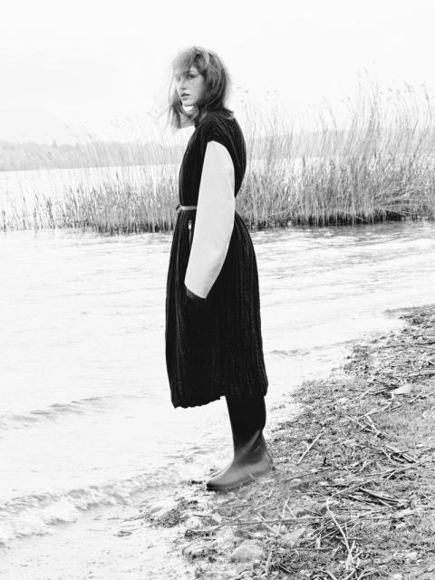 A black and white shot captures a woman in a dark dress and boots standing near water and tall grasses at the edge of a lake.