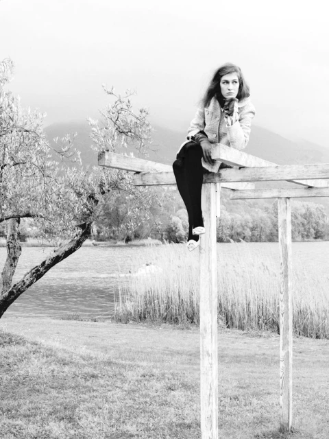 Black and white portrait of a young woman in a coat and scarf sitting on a wooden structure, with a lake and mountain in the background.