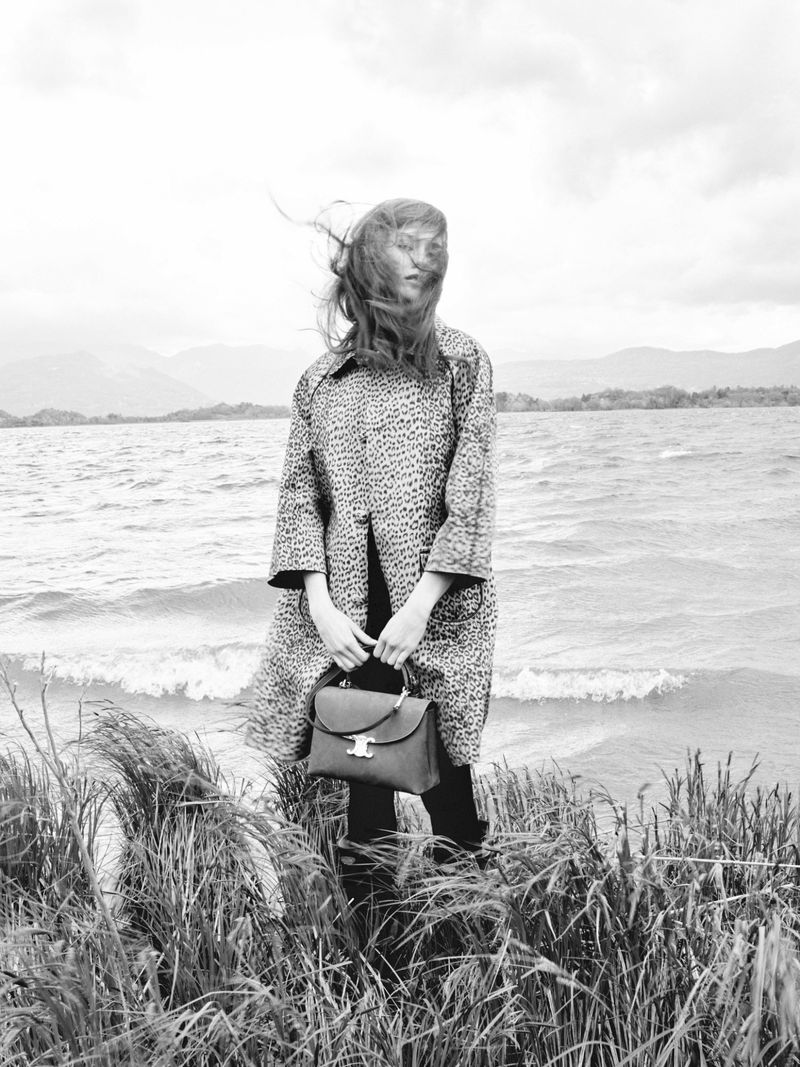 Monochrome portrait of a woman in a patterned coat holding a handbag by a lake with grassy foreground and distant mountains.