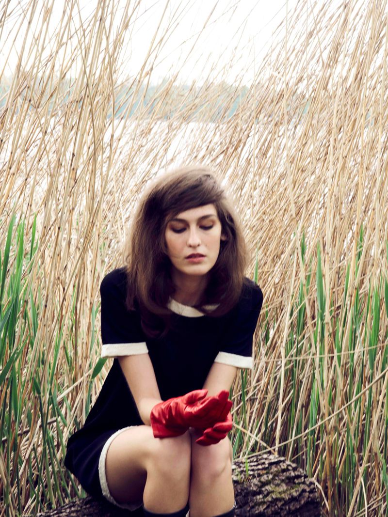 A woman in a black and white dress sits outdoors on a log in a field of tall grasses with a lake and trees in the background.