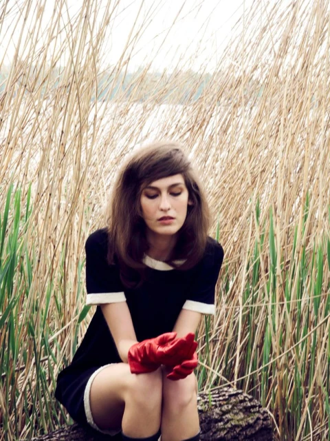 A woman in a black and white dress sits outdoors on a log in a field of tall grasses with a lake and trees in the background.