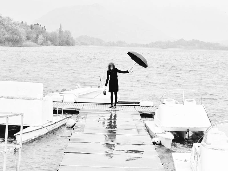 Monochrome photo of a woman on a dock holding an umbrella in the rain, surrounded by boats, with trees and mountains in the background.