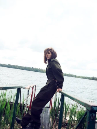 An adult female in a green jumpsuit poses on a metal bridge over a lake, featuring natural light, a full shot, and serene outdoor setting.
