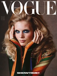 Close-up portrait of a woman with voluminous blonde hair and blue eyeshadow wearing a striped top against a dark studio background