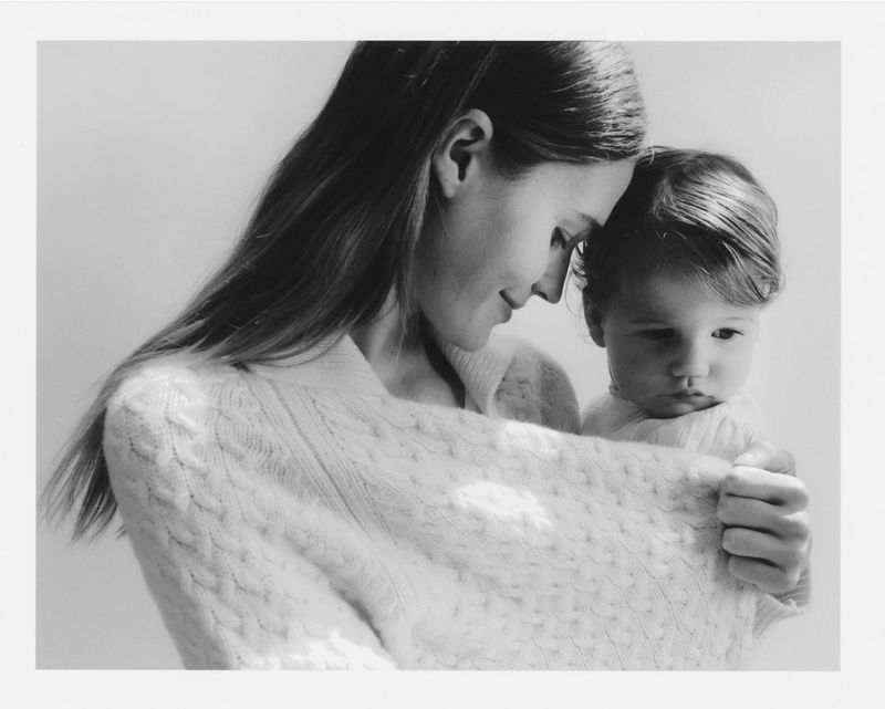A black and white photograph shows a mother and child, the mother looking down at the child with a gentle expression.