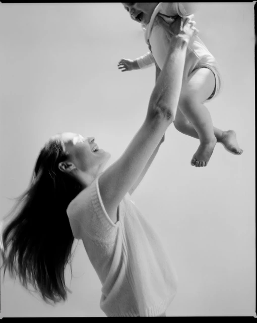 A happy adult female holding a laughing baby up in the air, both smiling; monochromatic shot with bright and airy background.