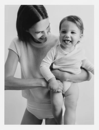 Monochrome portrait of a smiling adult woman holding a happy baby who is sticking out their tongue in front of a white backdrop.