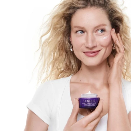 Woman applying cream to her cheek while holding a Clinique smart clinical repair jar against a white background