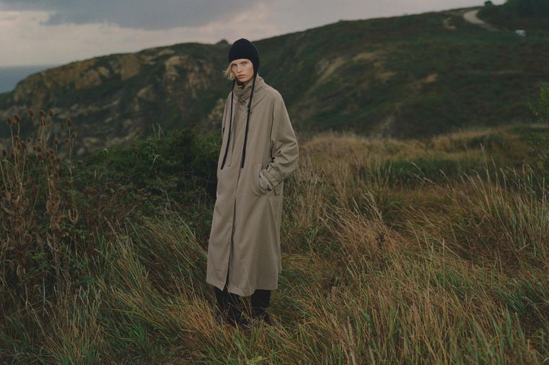 Model standing in a grassy coastal landscape wearing a long coat and black beanie for Massimo Dutti Raw & Untamed.