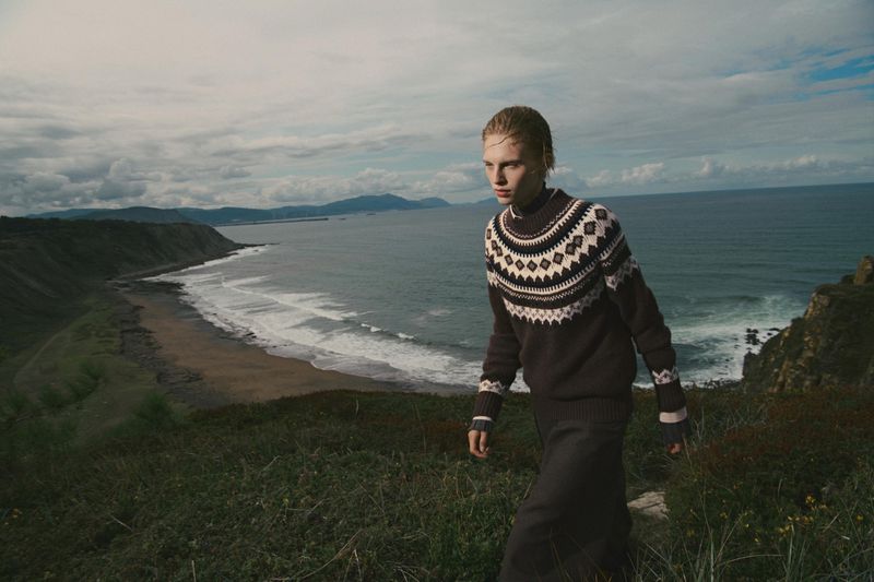 Model wearing a dark Fair Isle patterned sweater walking along a rugged coastal cliffside overlooking the ocean.