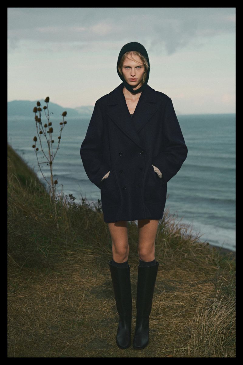 Model wearing a dark coat and hood standing on a coastal cliff above the ocean for Massimo Dutti