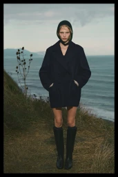 Model wearing a dark coat and hood standing on a coastal cliff above the ocean for Massimo Dutti