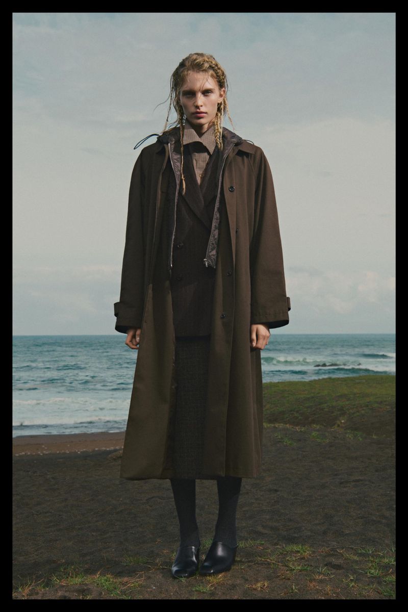 Model standing outdoors wearing a long brown coat and dark shoes with an ocean backdrop under an overcast sky.