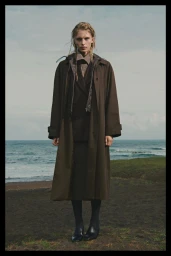 Model standing outdoors wearing a long brown coat and dark shoes with an ocean backdrop under an overcast sky.