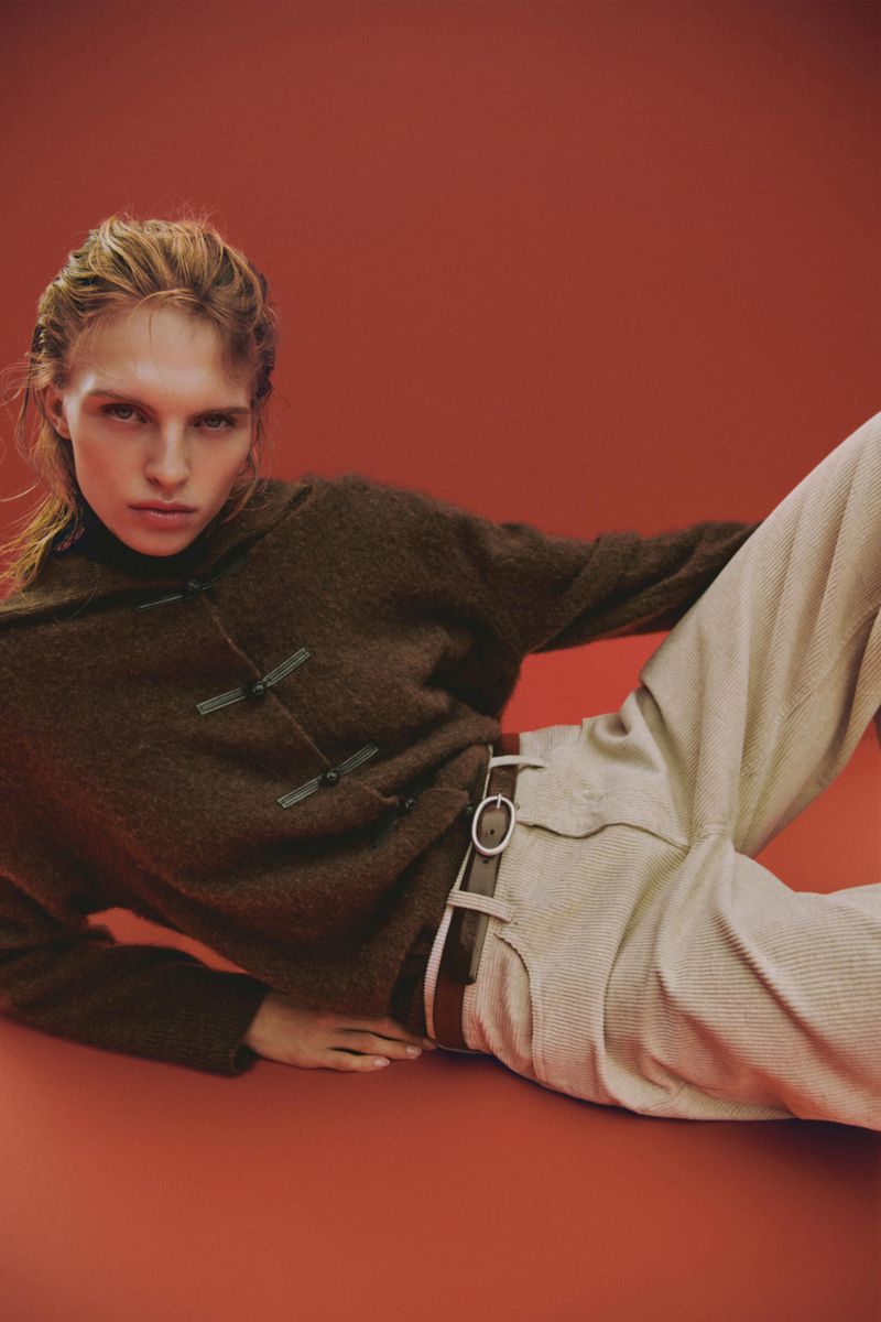 Model wearing a brown knit sweater and light trousers lying against a warm red studio background