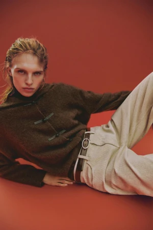 Model wearing a brown knit sweater and light trousers lying against a warm red studio background