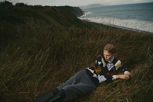 Model lying in tall grassy coastal dunes wearing a patterned sweater and dark skirt with ocean view