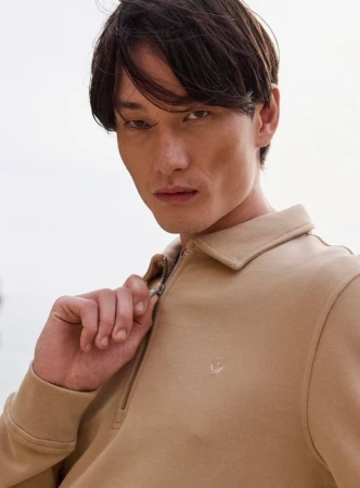 Man wearing a beige quarter-zip polo shirt holding the zipper against an overcast sky background.