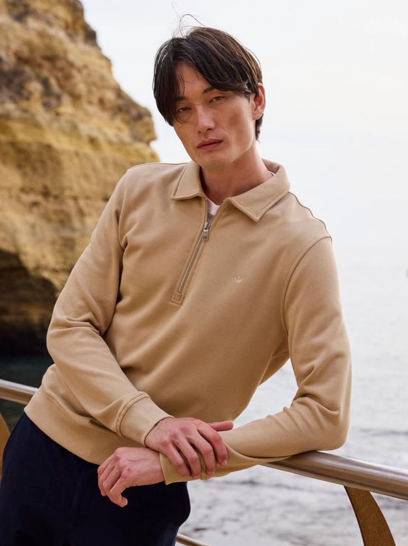 Model leaning on a railing against a coastal cliffside background wearing a beige half-zip sweater.