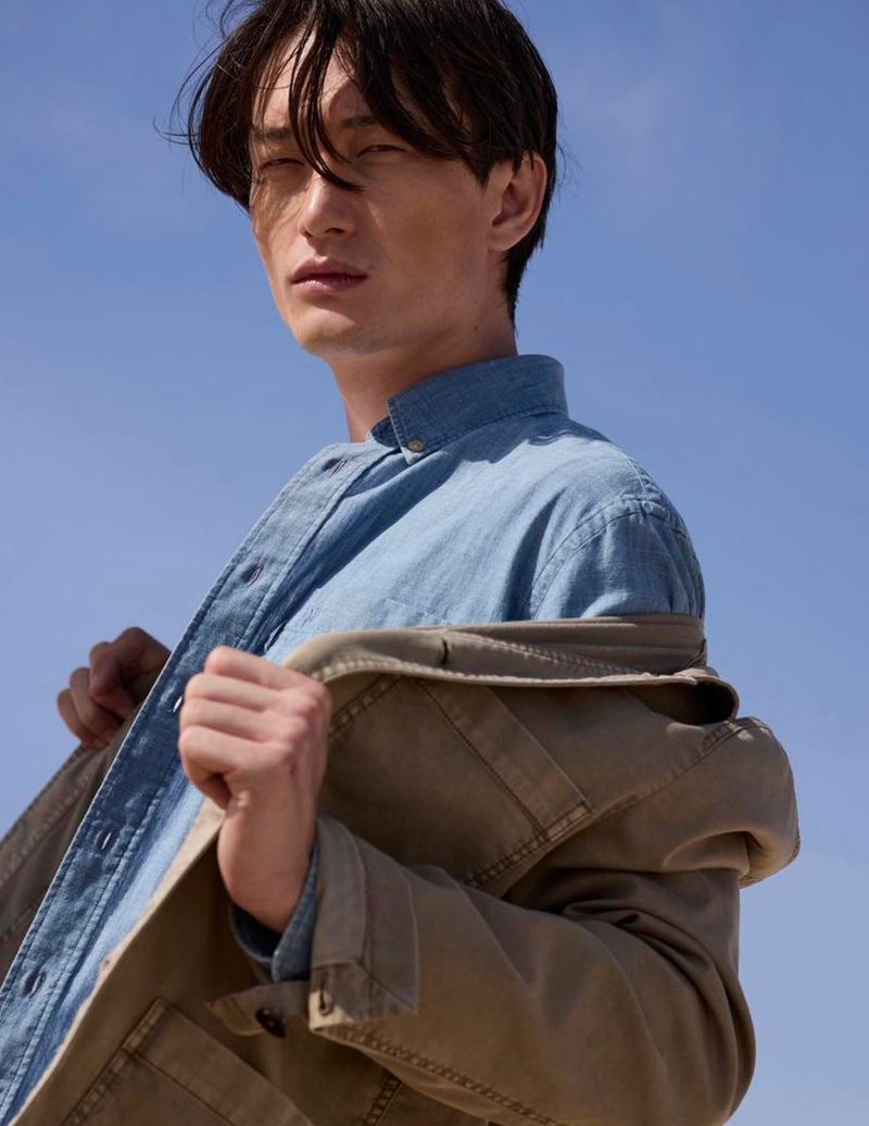 Low-angle shot of a male model in a denim shirt wearing a tan jacket against a clear blue sky background