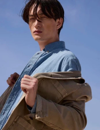 Low-angle shot of a male model in a denim shirt wearing a tan jacket against a clear blue sky background