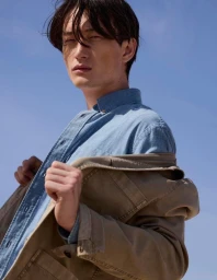 Low-angle shot of a male model in a denim shirt wearing a tan jacket against a clear blue sky background
