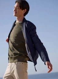 Man wearing a navy bomber jacket and olive sweater outdoors against a clear blue sky background