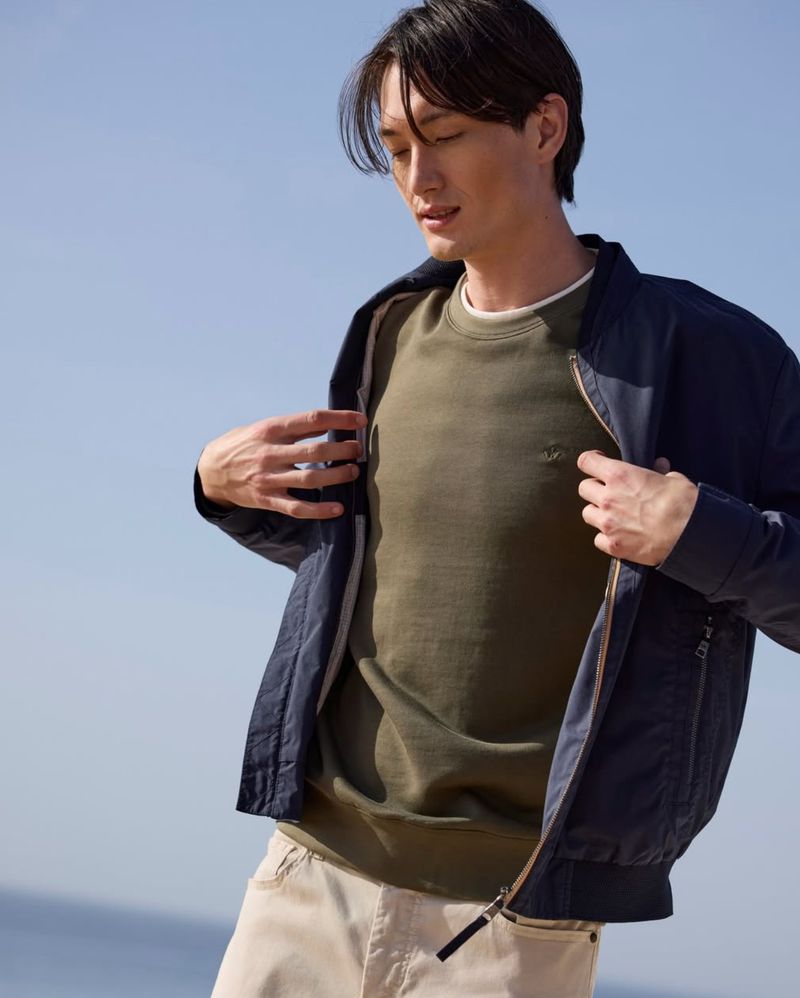 Man wearing an olive green sweatshirt and navy bomber jacket outdoors against a clear blue sky