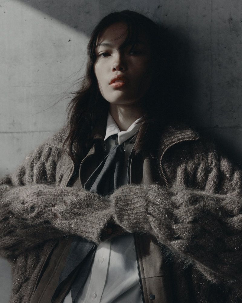 Model in a textured knit sweater and collared shirt posing against a concrete wall for Brunello Cucinelli FW26