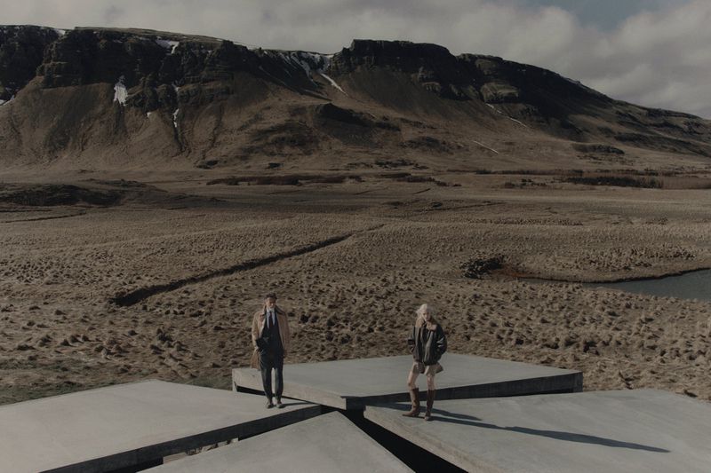Two models standing on concrete platforms in a vast rugged landscape with mountains under an overcast sky.