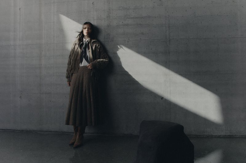 Model leaning against a concrete wall wearing a brown pleated skirt and textured jacket in dramatic light