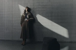 Model leaning against a concrete wall wearing a brown pleated skirt and textured jacket in dramatic light