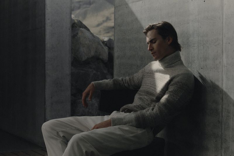 A man wearing a grey turtleneck sweater and white trousers seated against a concrete wall with a rocky outdoor backdrop.