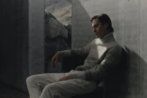 A man wearing a grey turtleneck sweater and white trousers seated against a concrete wall with a rocky outdoor backdrop.