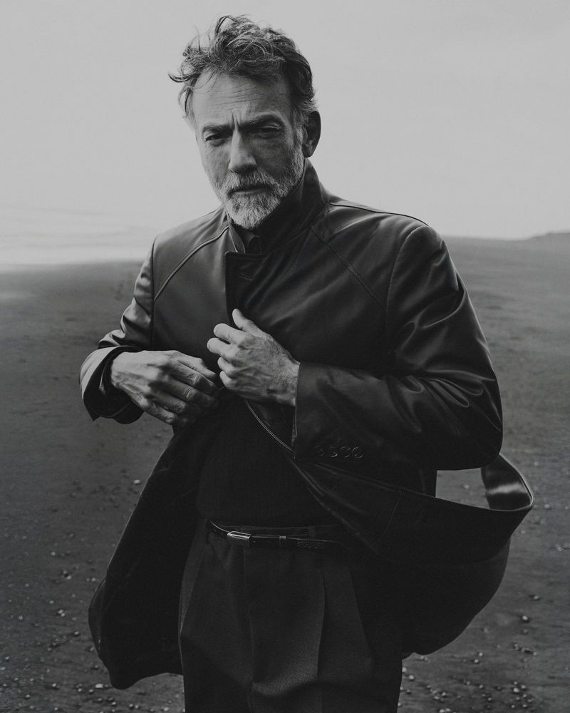 Monochrome portrait of a mature male model adjusting his dark leather coat against a sandy coast backdrop.