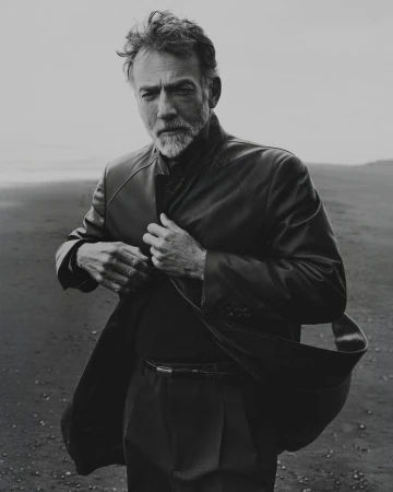 Monochrome portrait of a mature male model adjusting his dark leather coat against a sandy coast backdrop.