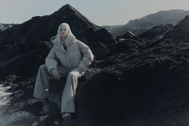 Model with platinum blonde hair seated on dark volcanic rocks wearing a padded puffer jacket and wide trousers
