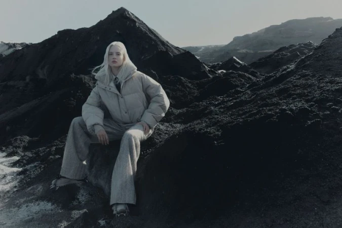 Model with platinum blonde hair seated on dark volcanic rocks wearing a padded puffer jacket and wide trousers