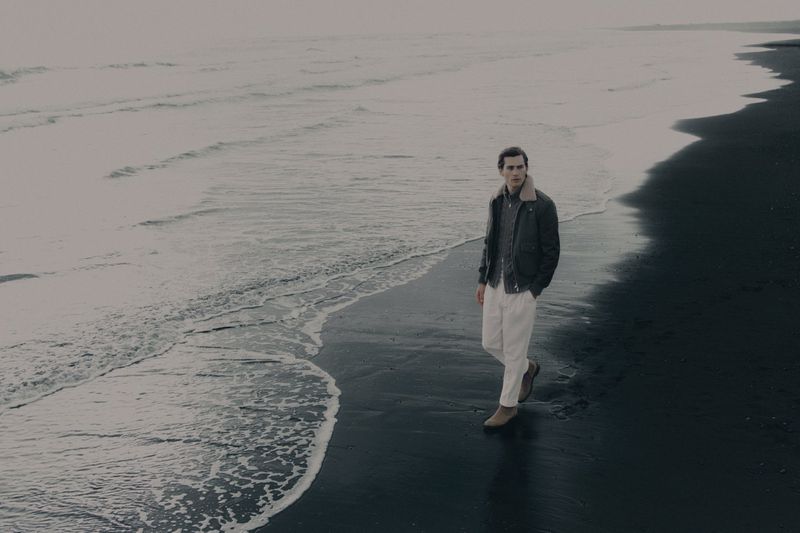 Man walking on a dark volcanic sandy beach wearing a dark jacket and white trousers