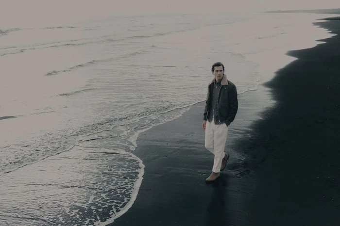 Man walking on a dark volcanic sandy beach wearing a dark jacket and white trousers