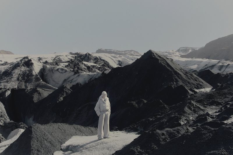 Model standing amid dark volcanic mountains and snowfields wearing winter outerwear for Brunello Cucinelli FW26 campaign.