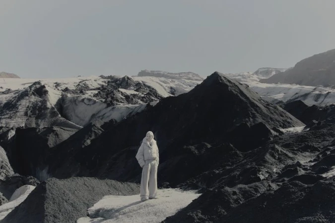 Model standing amid dark volcanic mountains and snowfields wearing winter outerwear for Brunello Cucinelli FW26 campaign.