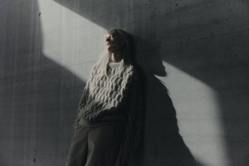 Model with blonde hair wearing a chunky cable-knit sweater leaning against a raw concrete wall in dramatic angular sunlight.