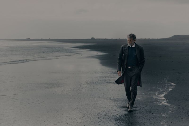 Man walking along a dark sand beach wearing a long dark coat for Brunello Cucinelli FW26