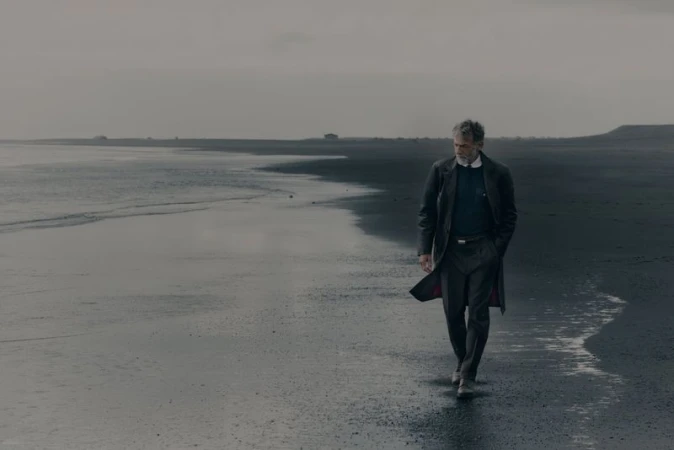 Man walking along a dark sand beach wearing a long dark coat for Brunello Cucinelli FW26