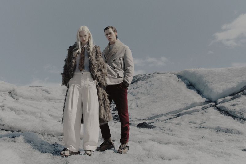 Two models standing on a snowy glacier wearing winter coats and trousers in an outdoor fashion campaign.