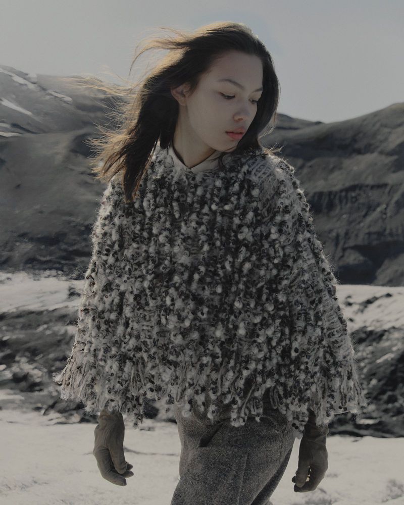 Model wearing a textured grey knit poncho and gloves outdoors against a snowy mountainous landscape under an overcast sky.